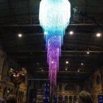 Photo of an electronically lit up chandelier, hanging in an event space, with people looking up at it in the foreground.