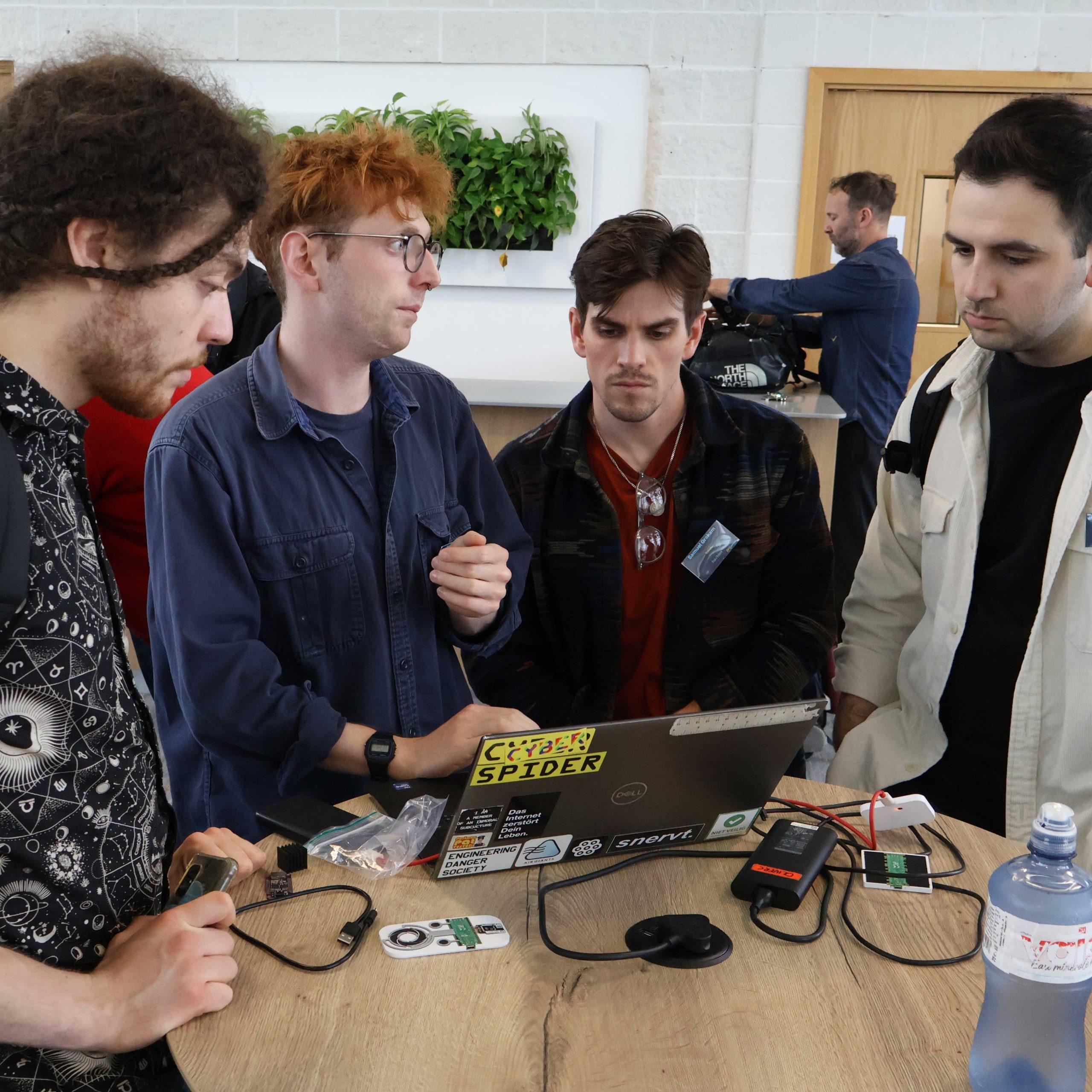 04 A group of four people stand around a small table looking at a laptop with electronic components beside it.