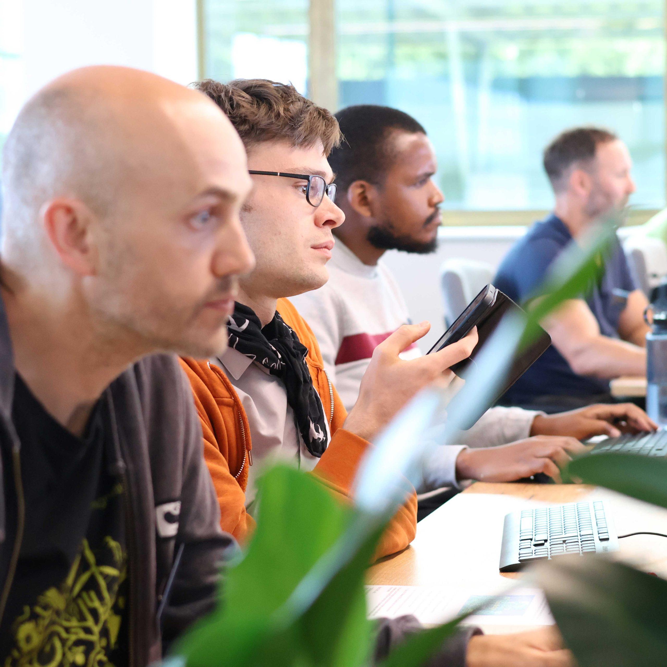 08 A row of four people sit in front of a desk in a computer lab with plants in the foreground.