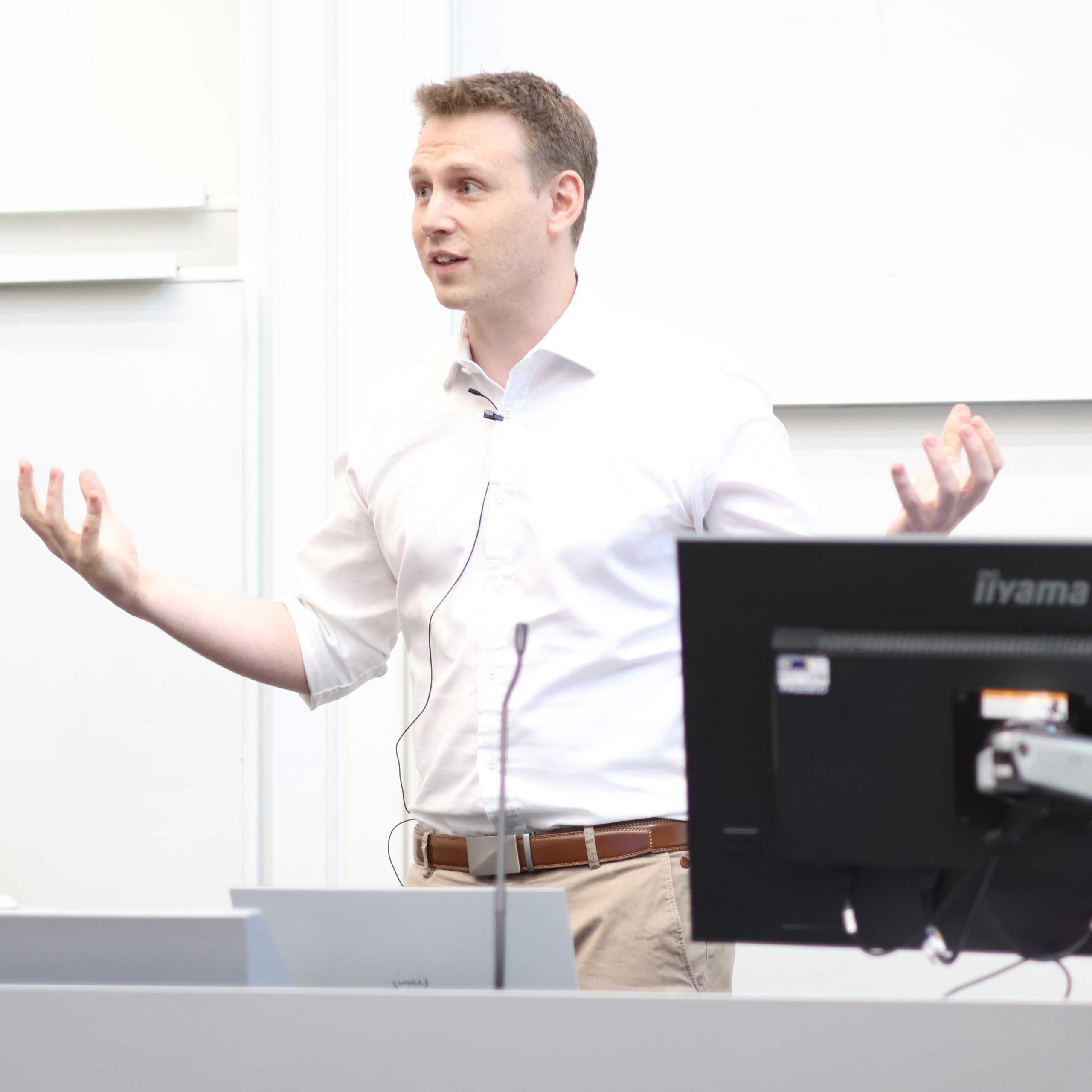 10 John Hardy gives a presentation at in a lecture theatre with a desk and monitor in the foreground.