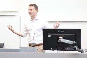 John Hardy stands in front of a desk with his hands raised to either side of him as he gives a talk at the summer school.