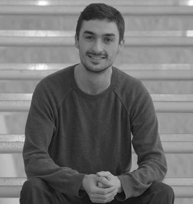 Kobi-Hartley A black and white photo of Kobi Hartley, sitting on a set of stairs with his hands clasped in front of him, smiling and facing the camera.