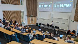 A group of people sit in a lecture theatre watching a person giving a talk. The presenter stands behind a desk with two large screens above showing the text 'Network Team' and photos.