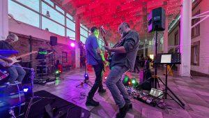 Three people play guitars at a concert with red and purple lighting and an audience in the background.