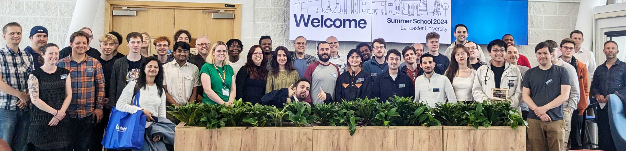 group A photo of the 2024 pro2 summer school attendees standing against a white brick wall, with plants in the foreground and a large screen reading 'Welcome, Summer School 2024, Lancaster University' and the pro2 logo in the background.