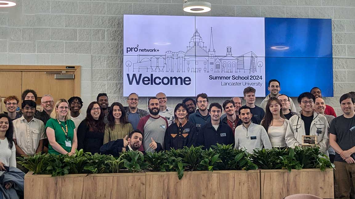 summer A photo of the 2024 pro2 summer school attendees standing against a white brick wall, with plants in the foreground and a large screen reading 'Welcome, Summer School 2024, Lancaster University' and the pro2 logo in the background.