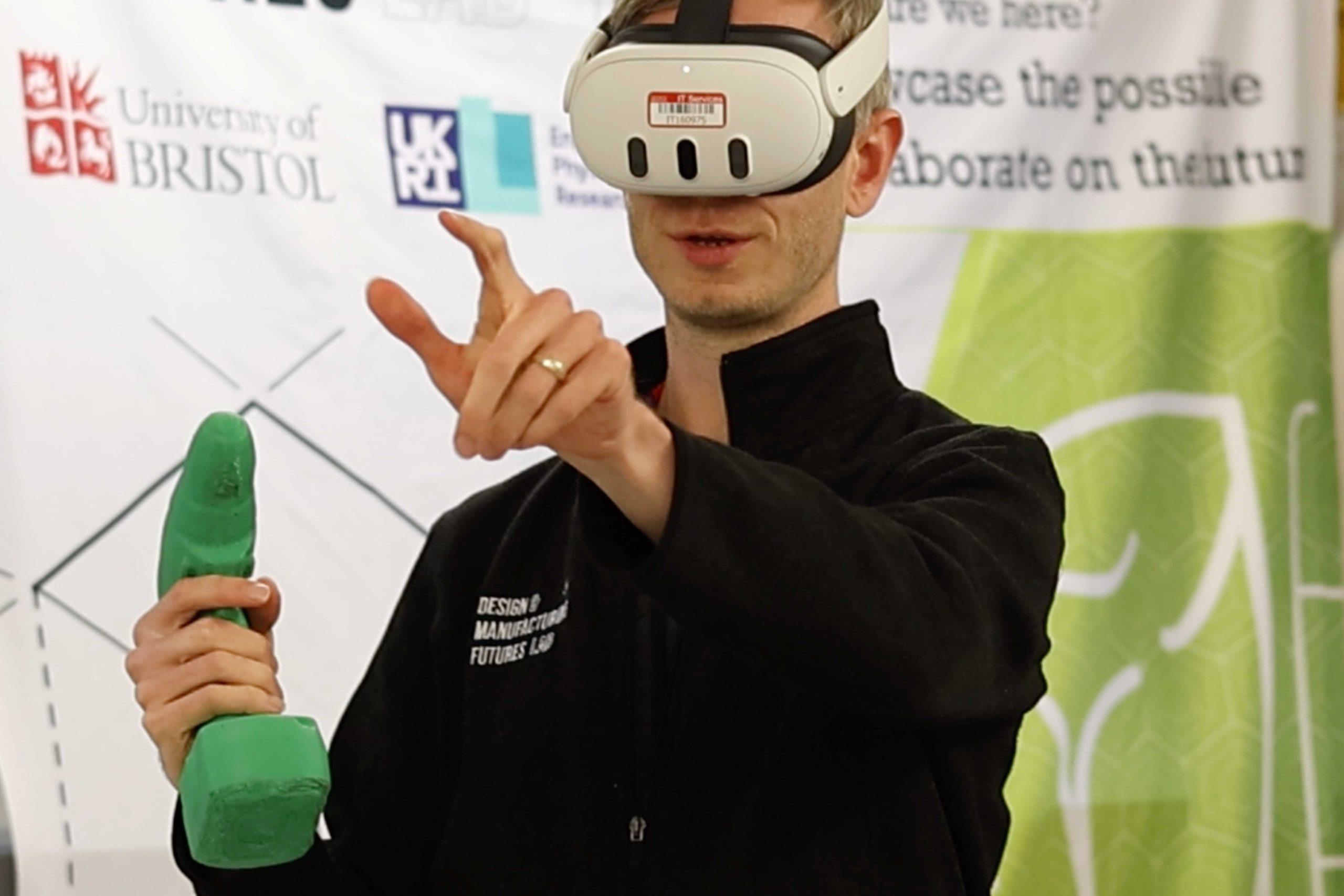 Multi-modal_01 A person standing in front of a banner with the University of Bristol logo, wearing a VR headset and holding a device prototype in one hand and pointing with the other.