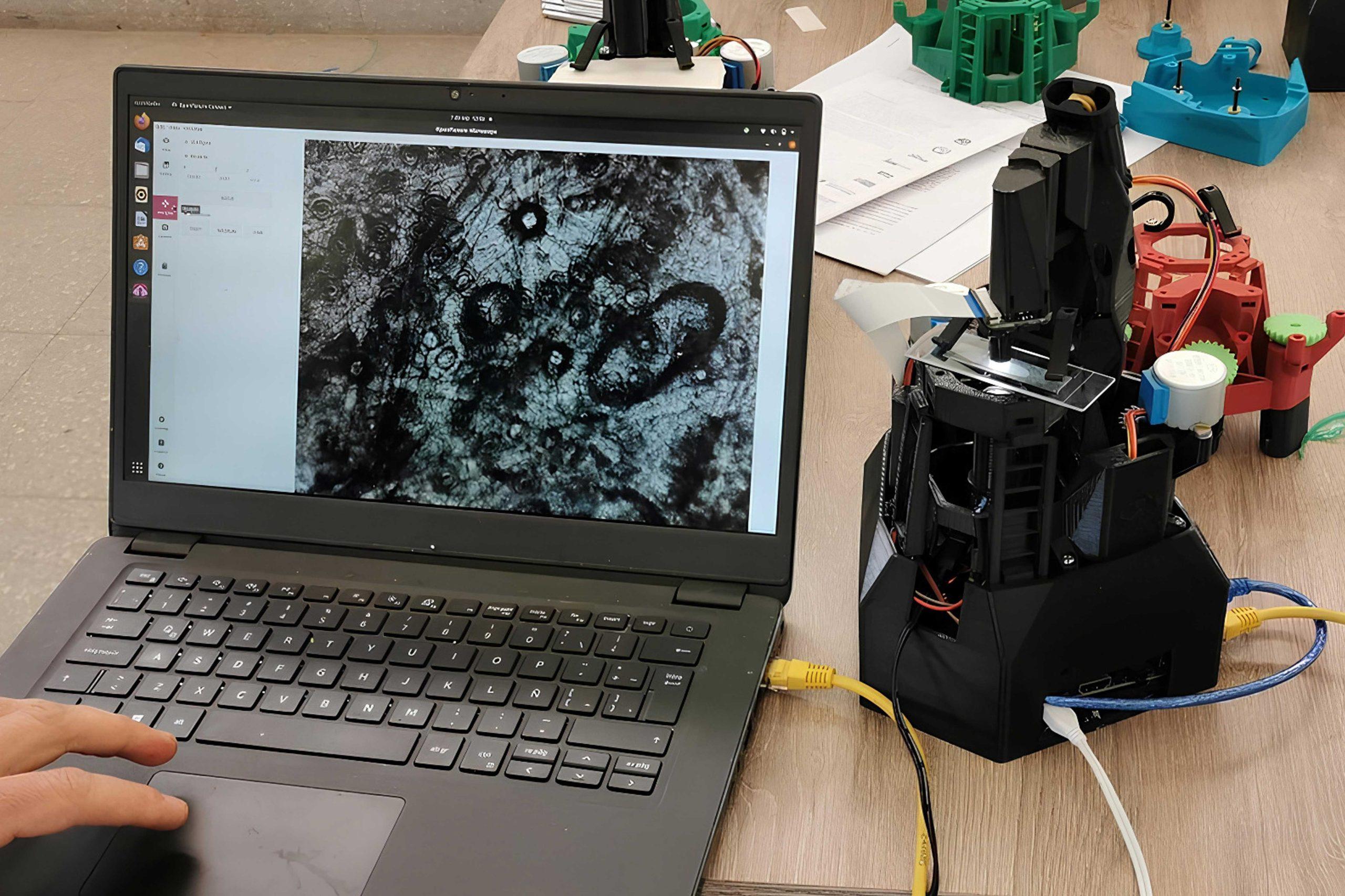 OpenFlexure_01 A photo of an open source microscope connected by a cable to a laptop. On the laptop screen is an image of the microscope sample.