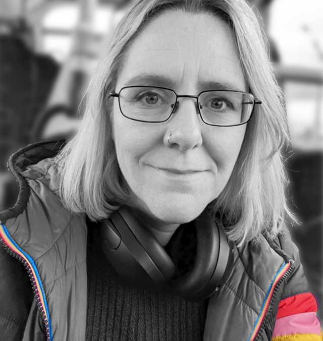 A black and white photo of Lorraine Underwood, smiling and looking toward the camera. She is wearing glasses, head phones around her neck, and a jacket with a small amount of colour on the sleeve.