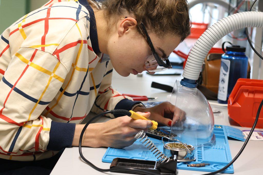 A 2024 summer school delegate leans over a desk in a lab while soldering a circuit board. She is wearing safety goggles and white, striped shirt.