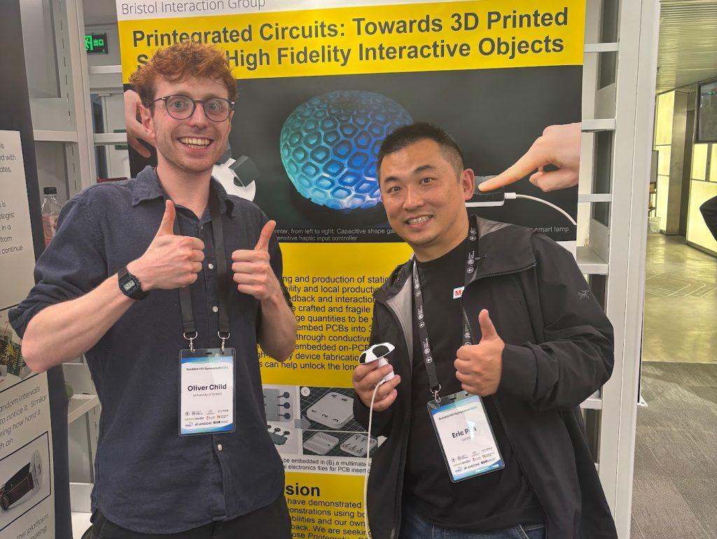 Oliver Child and Eric Pan standing in front of an academic poster. Both are smiling and giving thumbs up. Eric is holding a small mushroom shaped device prototype in one hand.