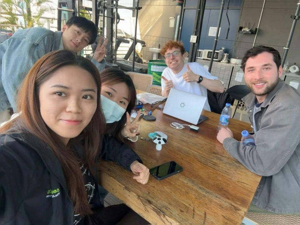 Oliver Child and his friend sit with a team of workers from Seeed Studio at the company's office. They are sitting around a wooden table with phones, a laptop and a device prototype.