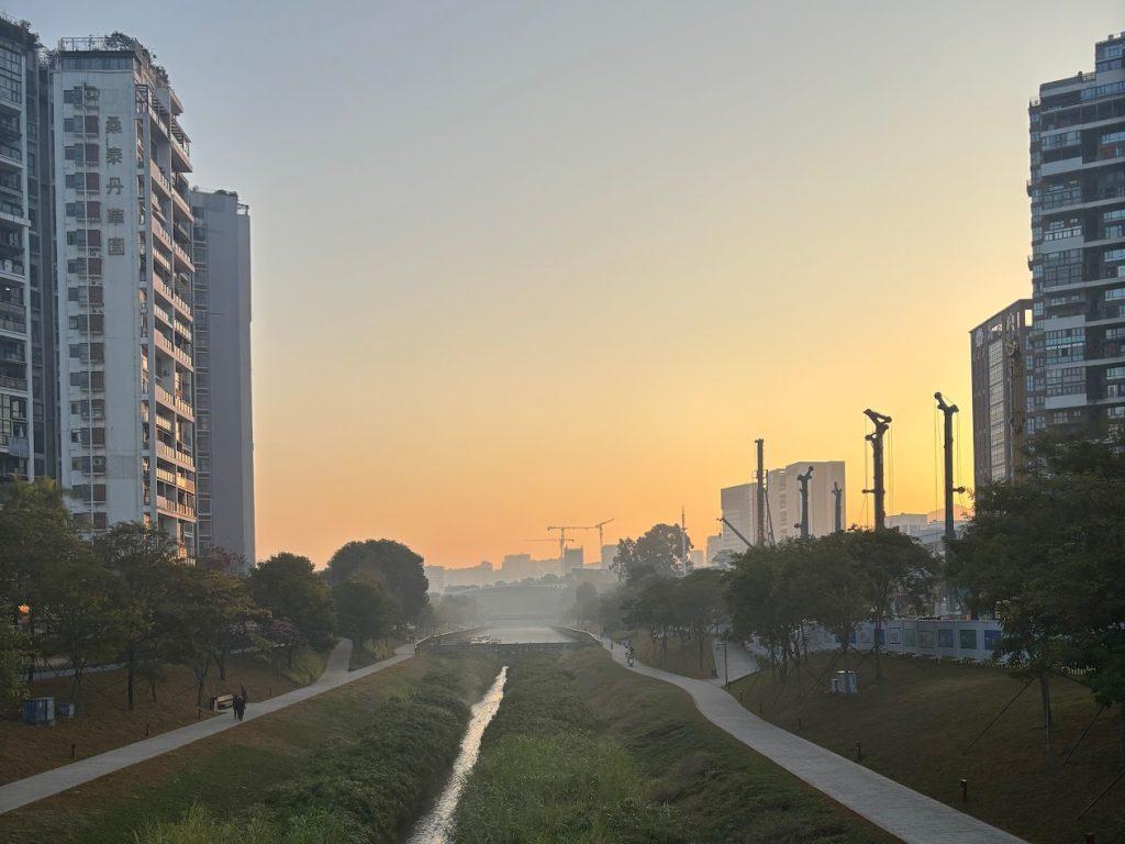 A photo of the city of Shenzhen at sunset. The sky glows orange and yellow, with tall buildings on either side of a river and construction cranes in the distance.