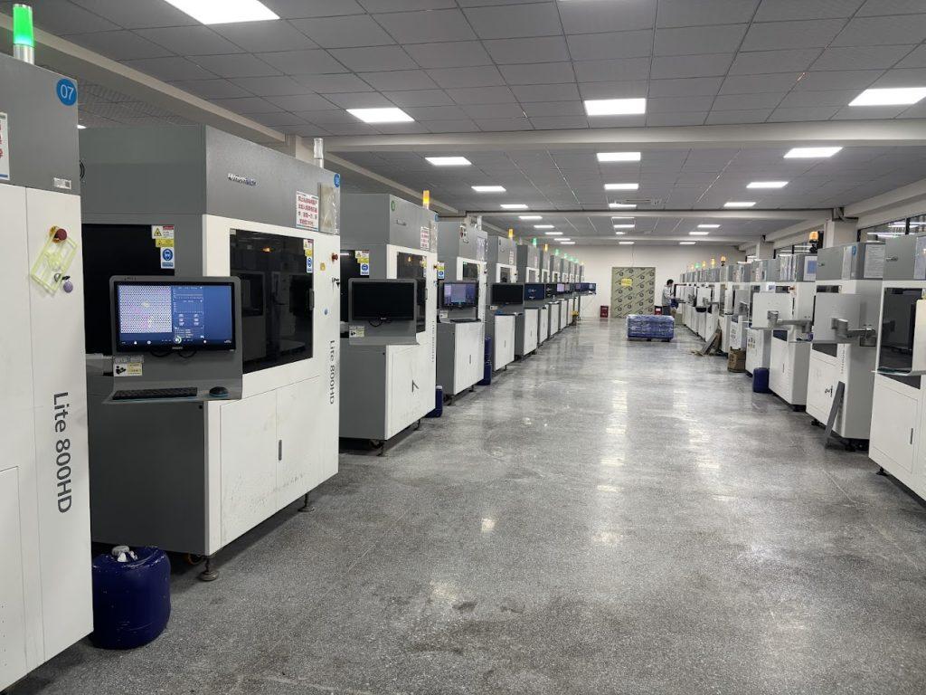 A large room in a factory full of 3D printers, with bright lights overhead and a worker in the distance.