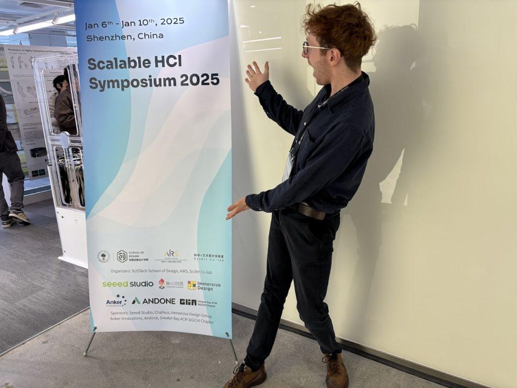 Oliver Child standing in front of a white wall, gesturing toward a long blue and white banner with text reading 'Scalable HCI Symposium 2025'.