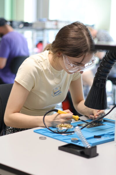 A summer school delegate holds a soldering iron while engaging in a hands-on surface mount assembly session in a lab.