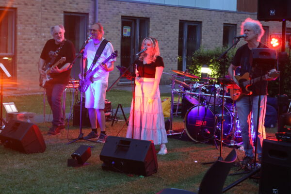 The band 'The Usuals' plays outside at night on the Lancaster University campus with concert lighting in the background.
