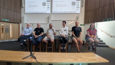 A panel of speakers presents in a lecture theatre with two screens displaying questions in the background.